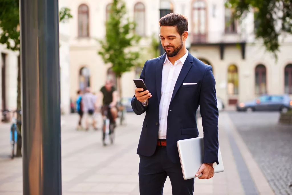 A suited man is holding a laptop and checking his phone, appearing engaged in work or communication.